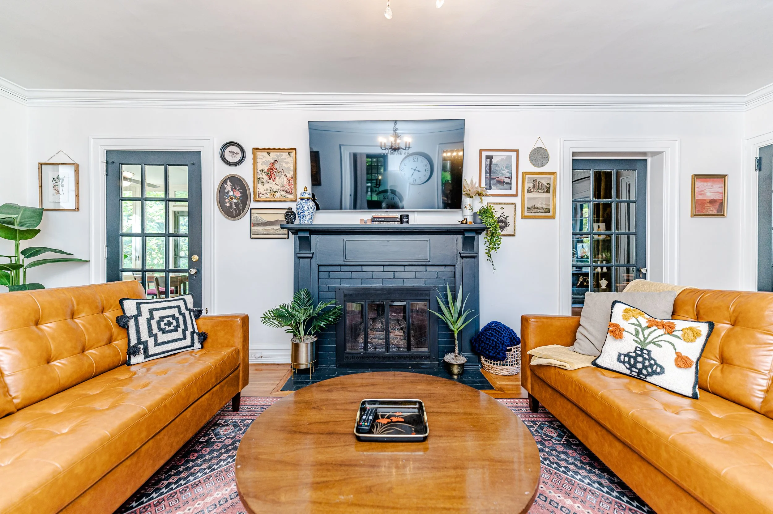 Living room with two tan leather sofas, a dark blue fireplace, wall art including paintings and framed pictures, a large flat-screen TV above the fireplace, and a wooden coffee table with a tray on it. Decor includes plants, pillows, a woven basket, and a patterned rug.