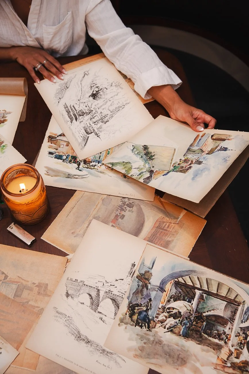 Person in a white shirt looking at open sketchbooks and watercolor paintings of cityscapes on a wooden table. A lit candle in an orange holder is beside the artwork.