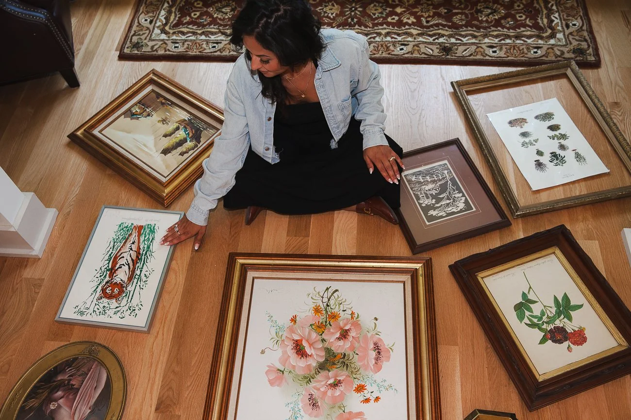 A woman sitting on the floor in a room with hardwood floors, surrounded by framed artwork laid on the floor.