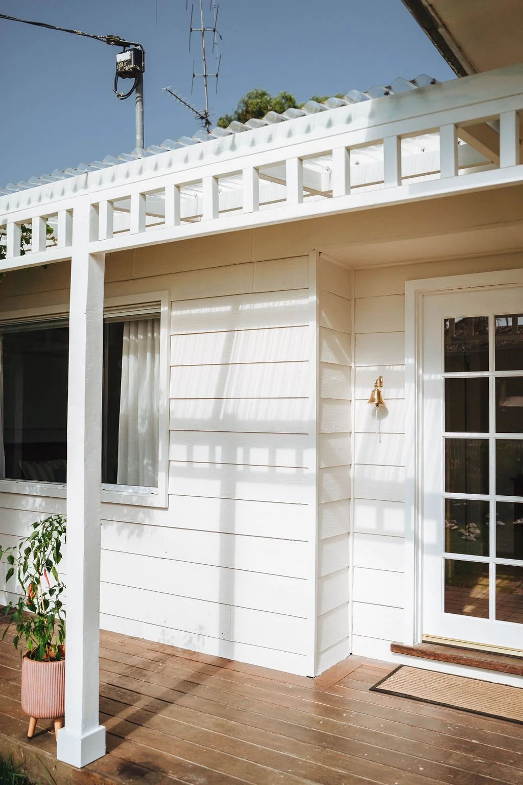 Backyard patio with white wooden siding, sliding glass door, potted plant, and wooden deck, under a bright blue sky.