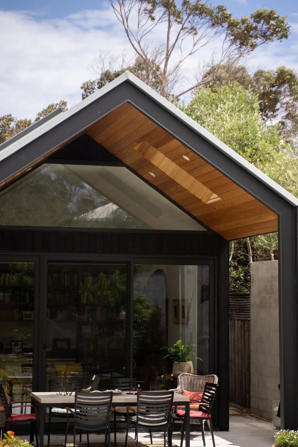 Exterior view of a modern house with a black metal frame, large glass sliding doors, and a wooden ceiling over the outdoor dining area, surrounded by trees and blue sky.