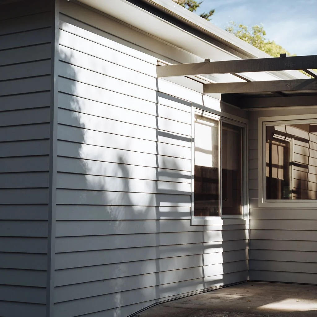 Close-up view of a house exterior with white horizontal siding, two windows with reflective glass, and an unfinished porch area with a wood beam protruding from the building.