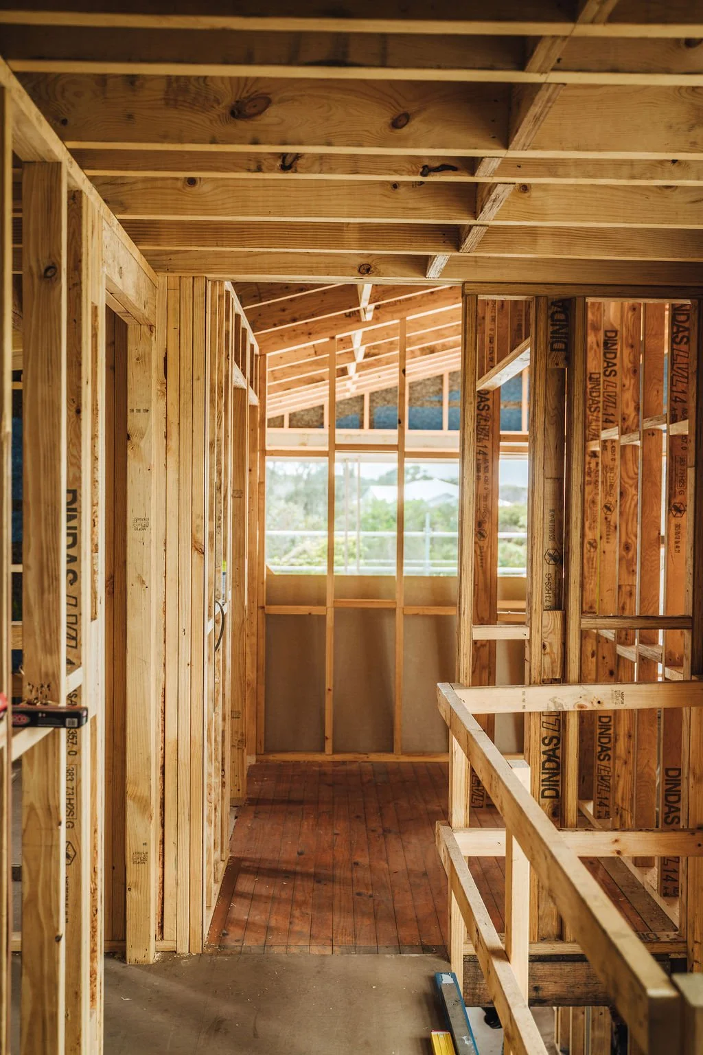 Interior view of a house under construction with exposed wooden framing, including walls, ceiling, and a partially built railing, with a window revealing an outdoor view.