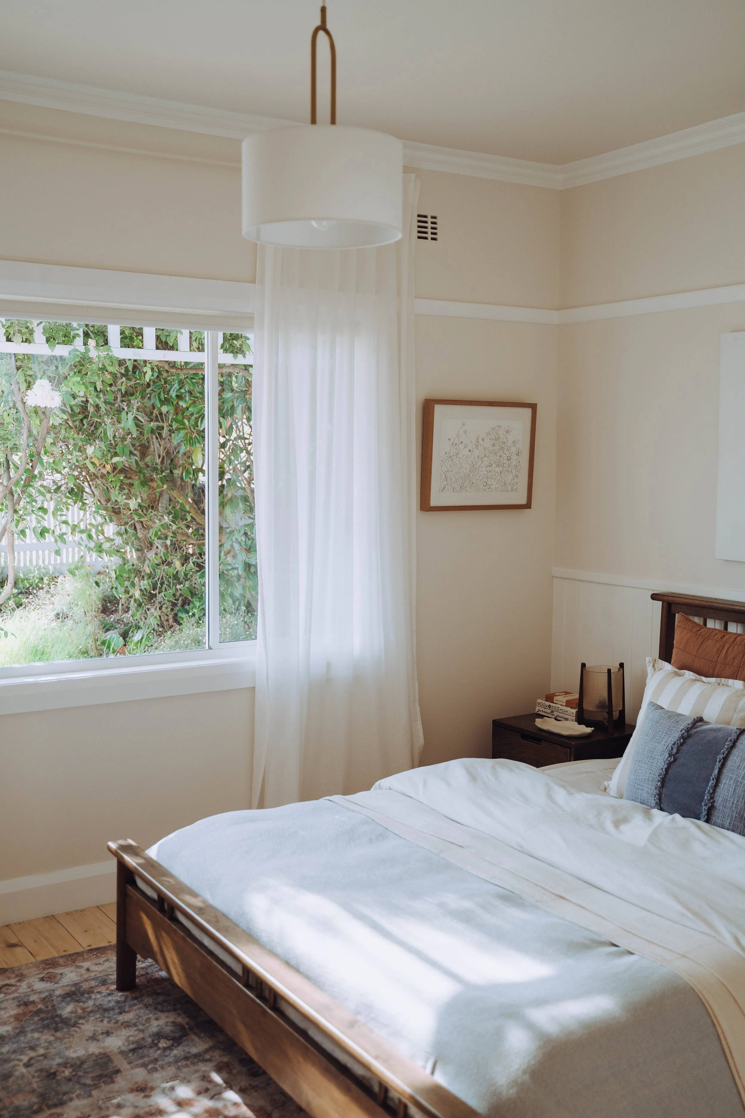 A cozy bedroom with a wooden bed frame, a white bedspread, and pillows. There's a window with white curtains revealing greenery outside. A framed botanical print hangs on the wall. A small nightstand holds books and decorative items. The room has light-colored walls and crown molding.