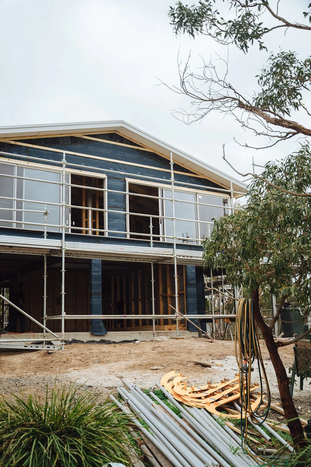 Construction site of a modern house with scaffolding, partially built exterior walls, and construction materials on the ground. Trees are visible on the right side.