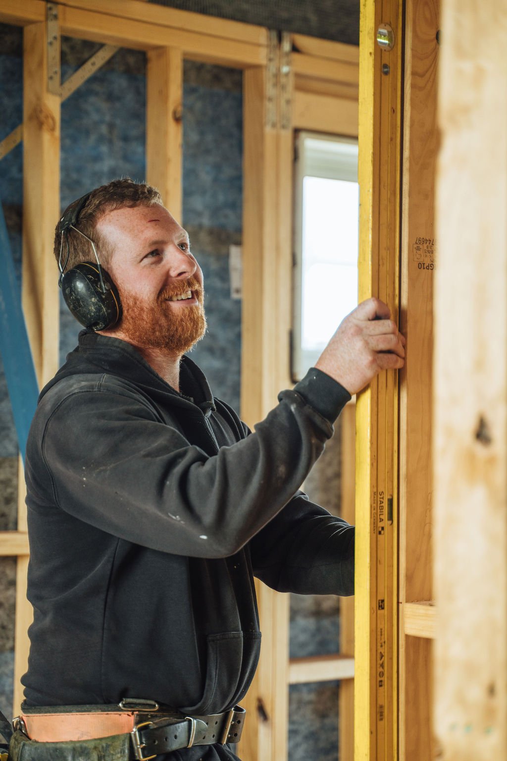 A man with a beard smiling while working on a wooden frame, wearing headphones and a dark jacket in an indoor construction site.