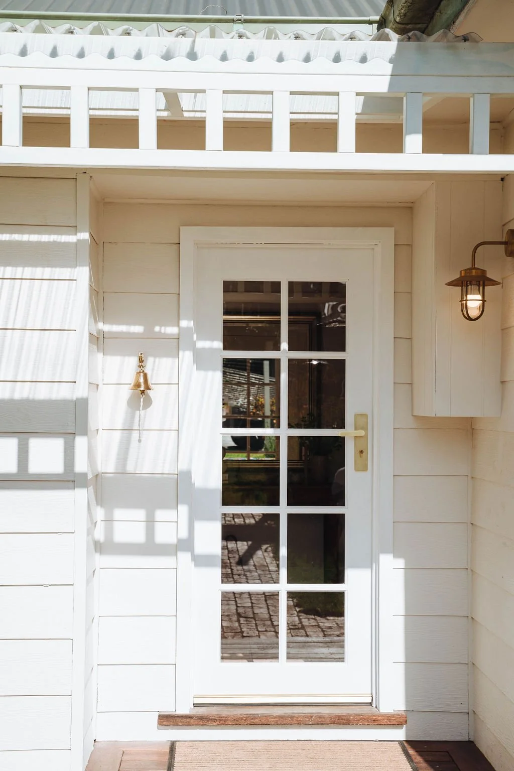 White door with glass panes on a light-colored wooden house exterior, flanked by wall-mounted light fixtures, leading to a porch with a doormat.