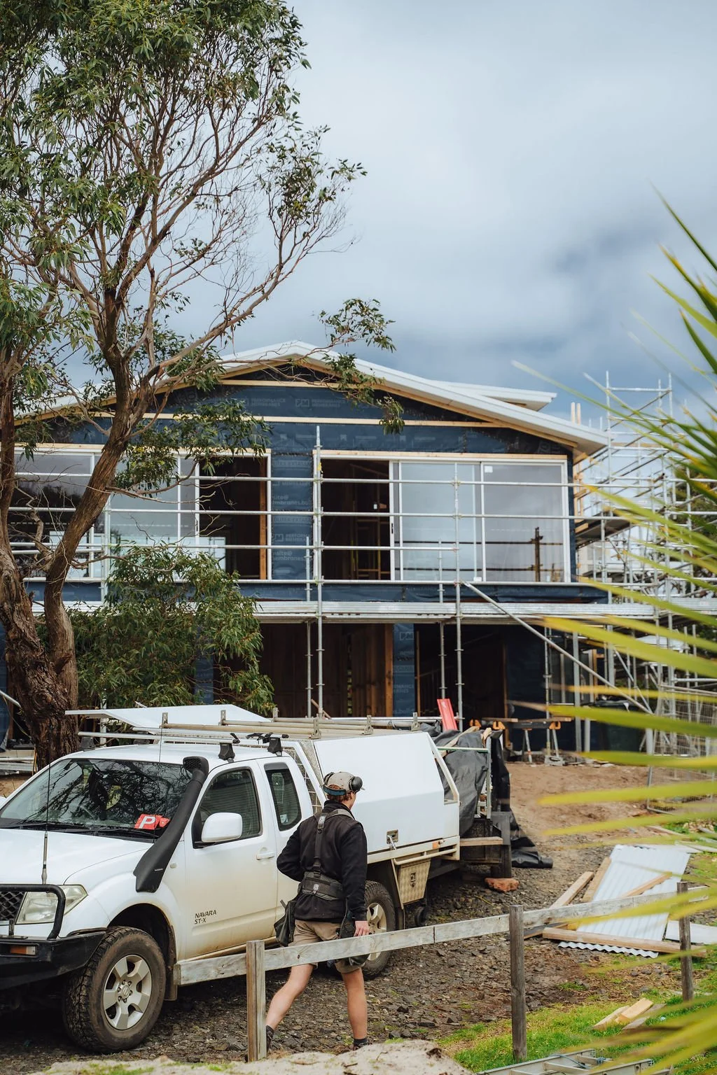 Construction site of a two-story house with scaffolding, a white utility truck parked in front, and a person in black clothing and a helmet walking nearby. The house has some exterior walls installed, but windows and other details are still in progress. A tree is in the foreground, and the sky is cloudy.
