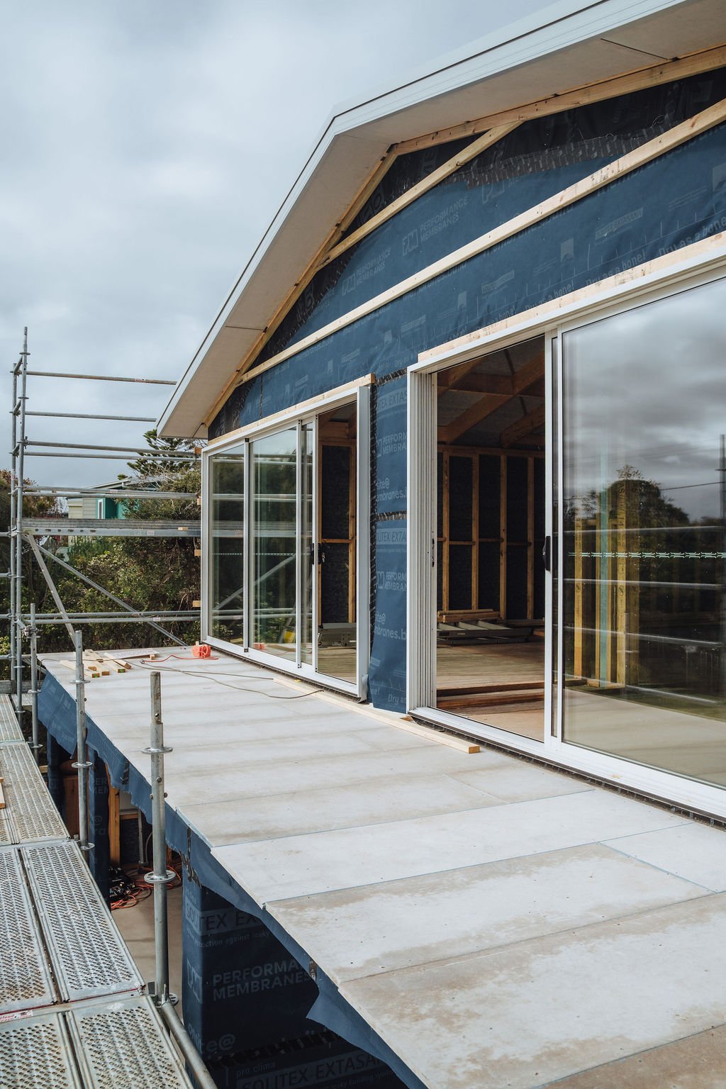 Construction of a house with large sliding glass doors, scaffolding on the side, and a sloped roof.