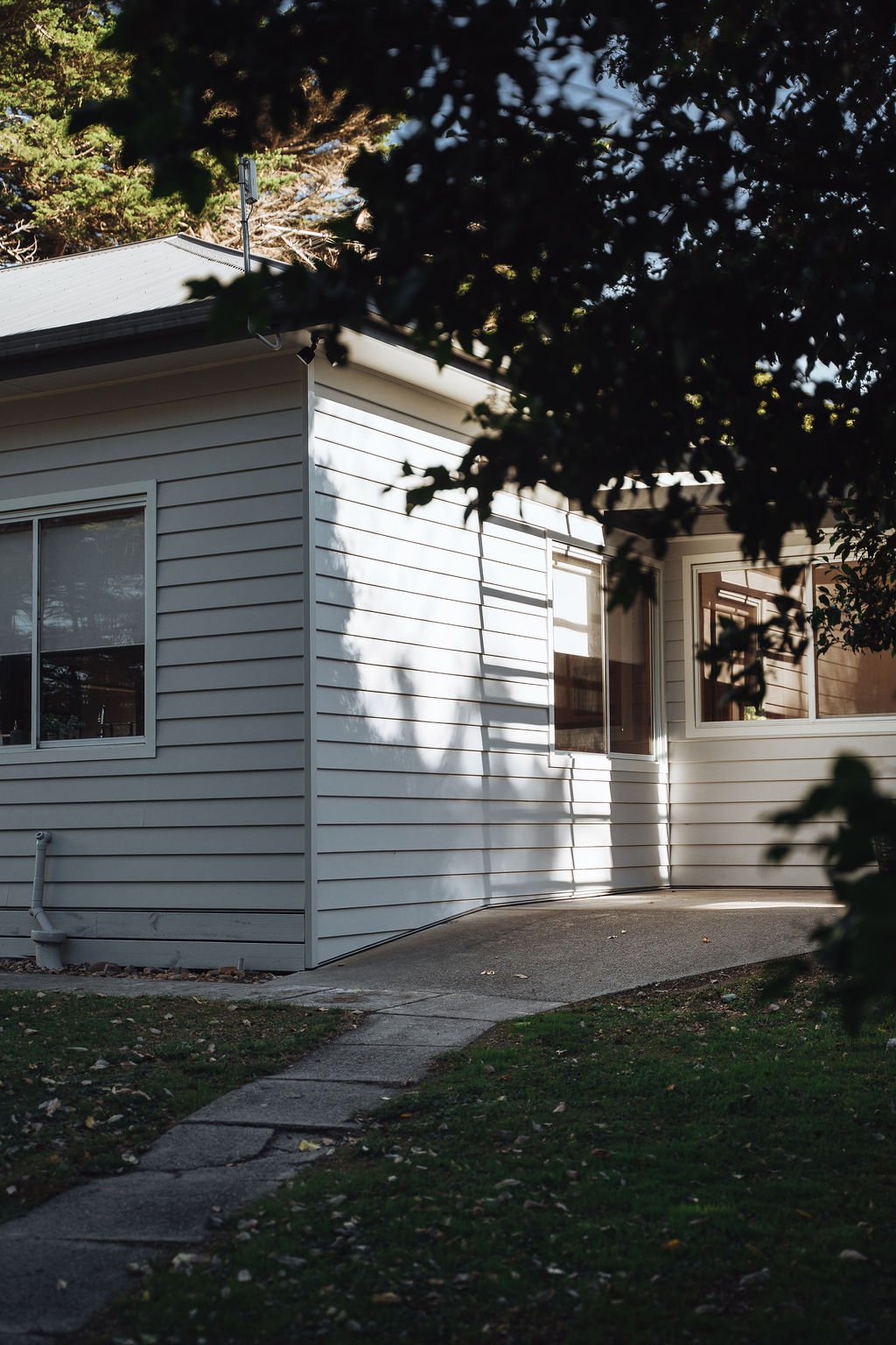 The side of a house with white siding, two windows, a small concrete pathway, and trees surrounding it, with shadows cast on the house.