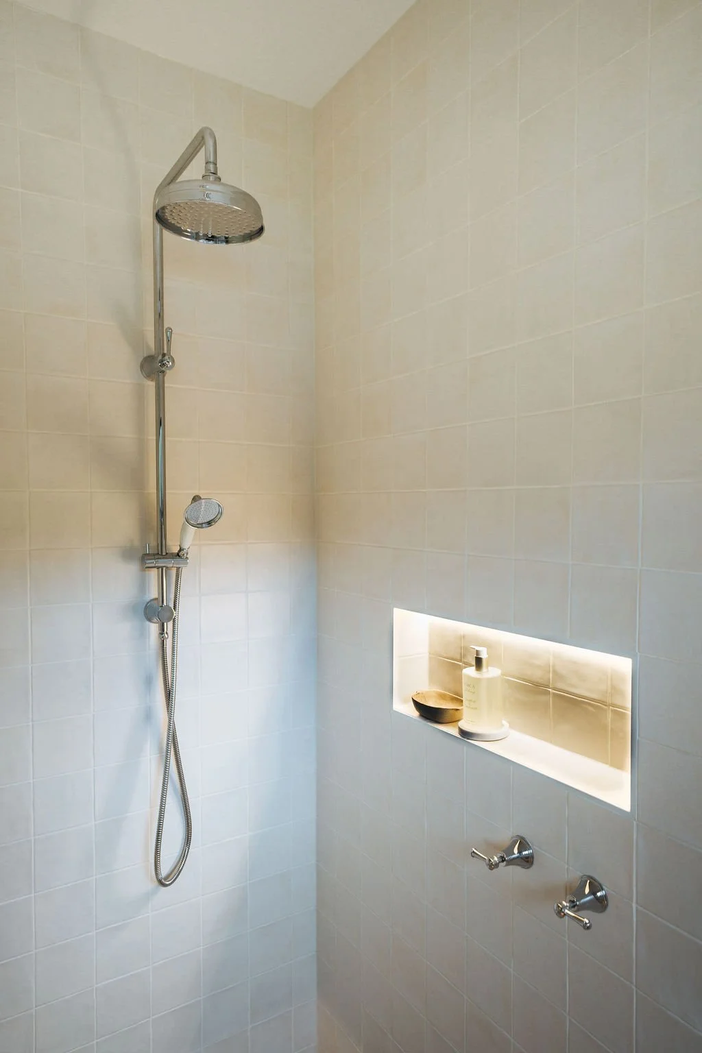 Shower area with a wall-mounted showerhead, a handheld shower wand, a niche with a soap dish and a bottle of body wash, and two water control knobs.