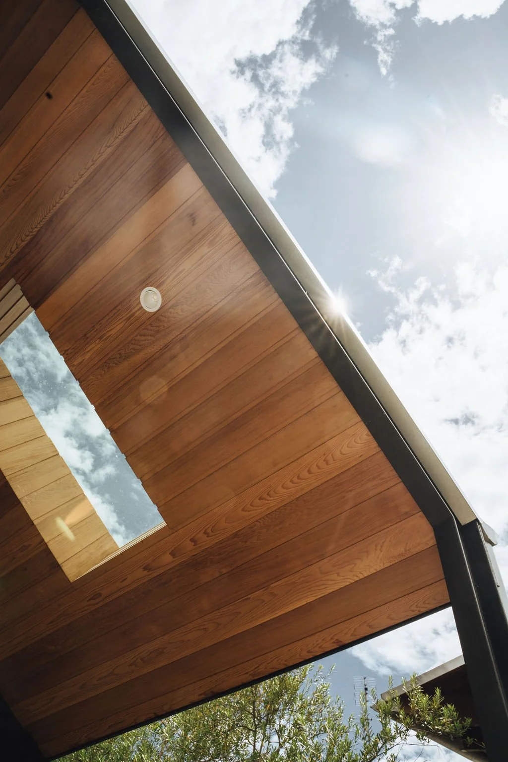 View of a modern building's wooden roof with a skylight, under a partly cloudy sky with sunlight visible.
