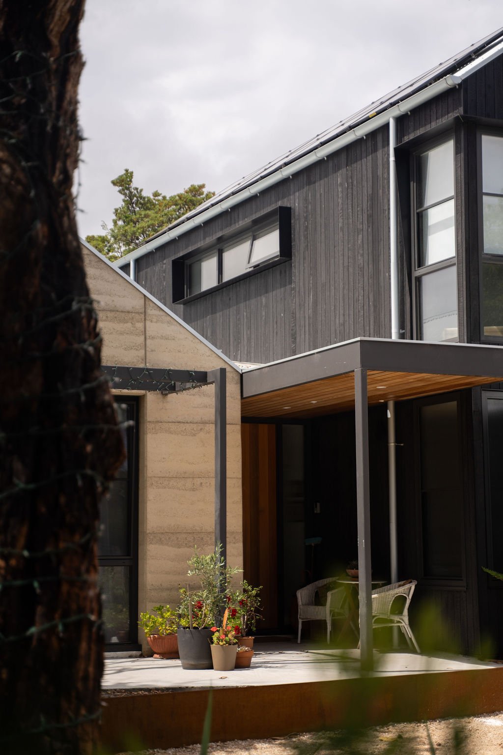 Modern house exterior with black wooden siding, large glass windows, a small porch with potted plants, and outdoor furniture.