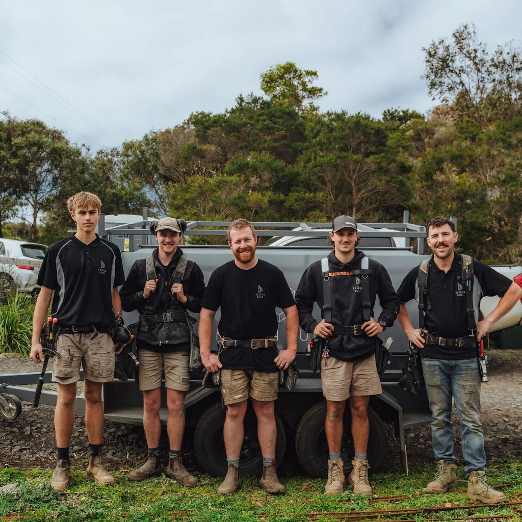 Five men standing outdoors in front of a utility trailer on a grassy area, with trees and cars in the background. They are dressed in outdoor work gear, mostly in black shirts and khaki shorts or pants, with some wearing backpacks and tools.