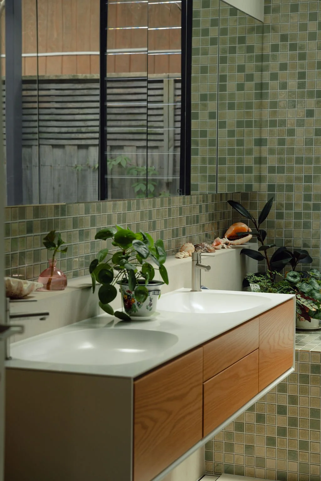 A bathroom with green mosaic tiles, a large mirror, and a modern wooden vanity with two sinks. The vanity is decorated with potted plants, seashells, and a small vase. Outside the window, a wooden fence and greenery are visible.