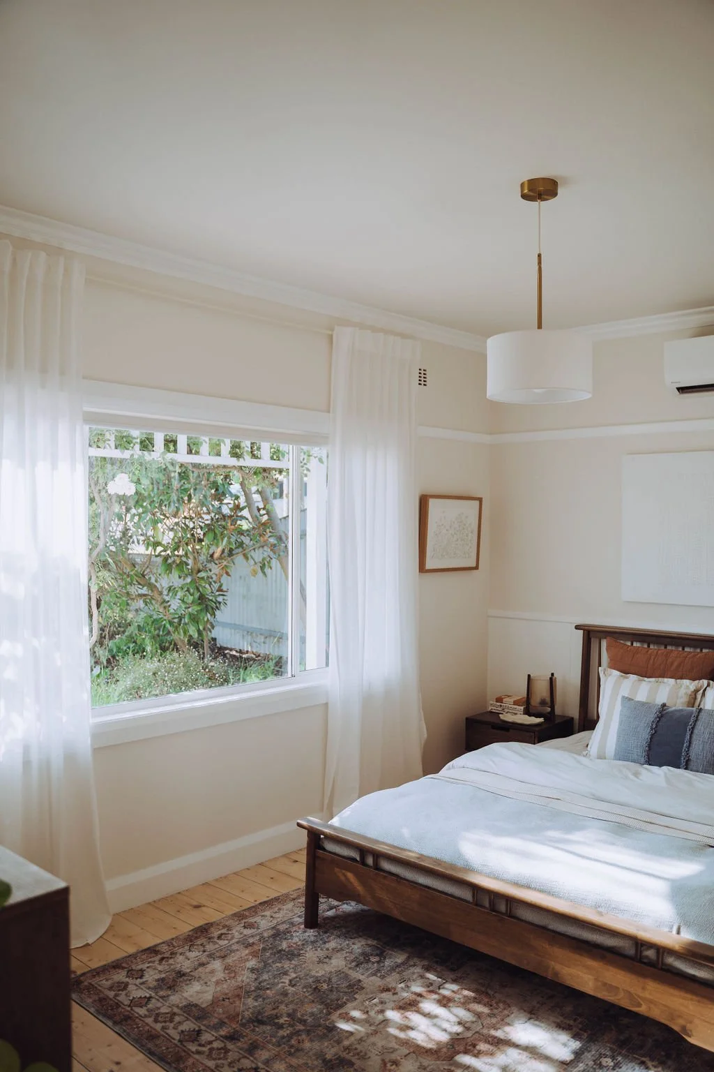 Bright bedroom with a large window, white curtains, wooden bed frame, navy and white striped pillows, nightstand, framed artwork on wall, pendant light, and air conditioning unit.