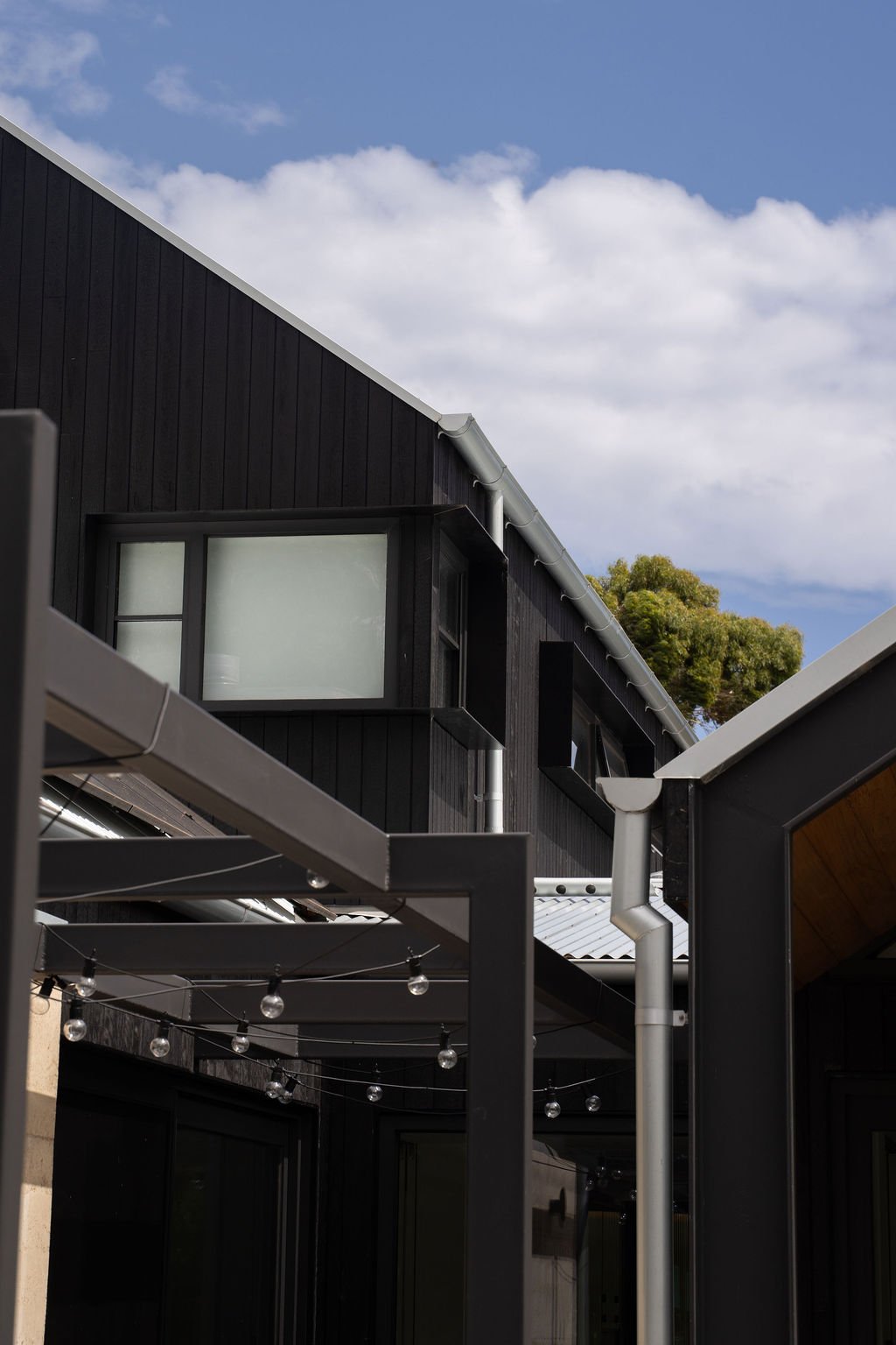 Modern black residential building with large windows, gray gutter system, and string lights under a partly cloudy sky.