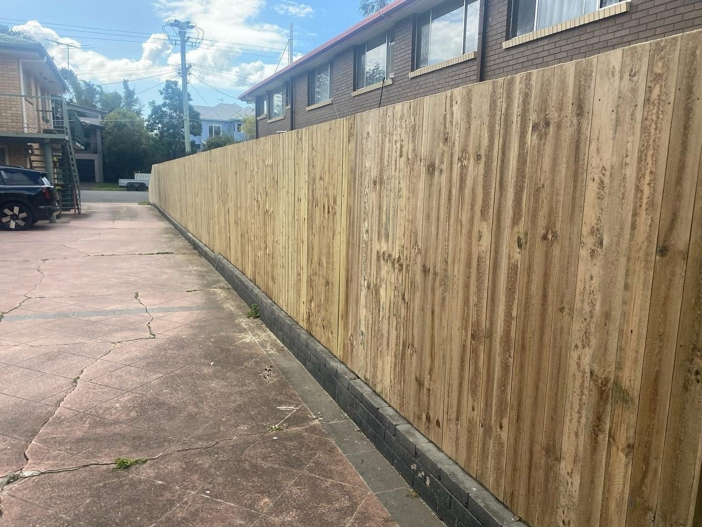 A newly installed wooden privacy fence along a concrete parking area on a sunny day. Residential buildings and cars are visible in the background.