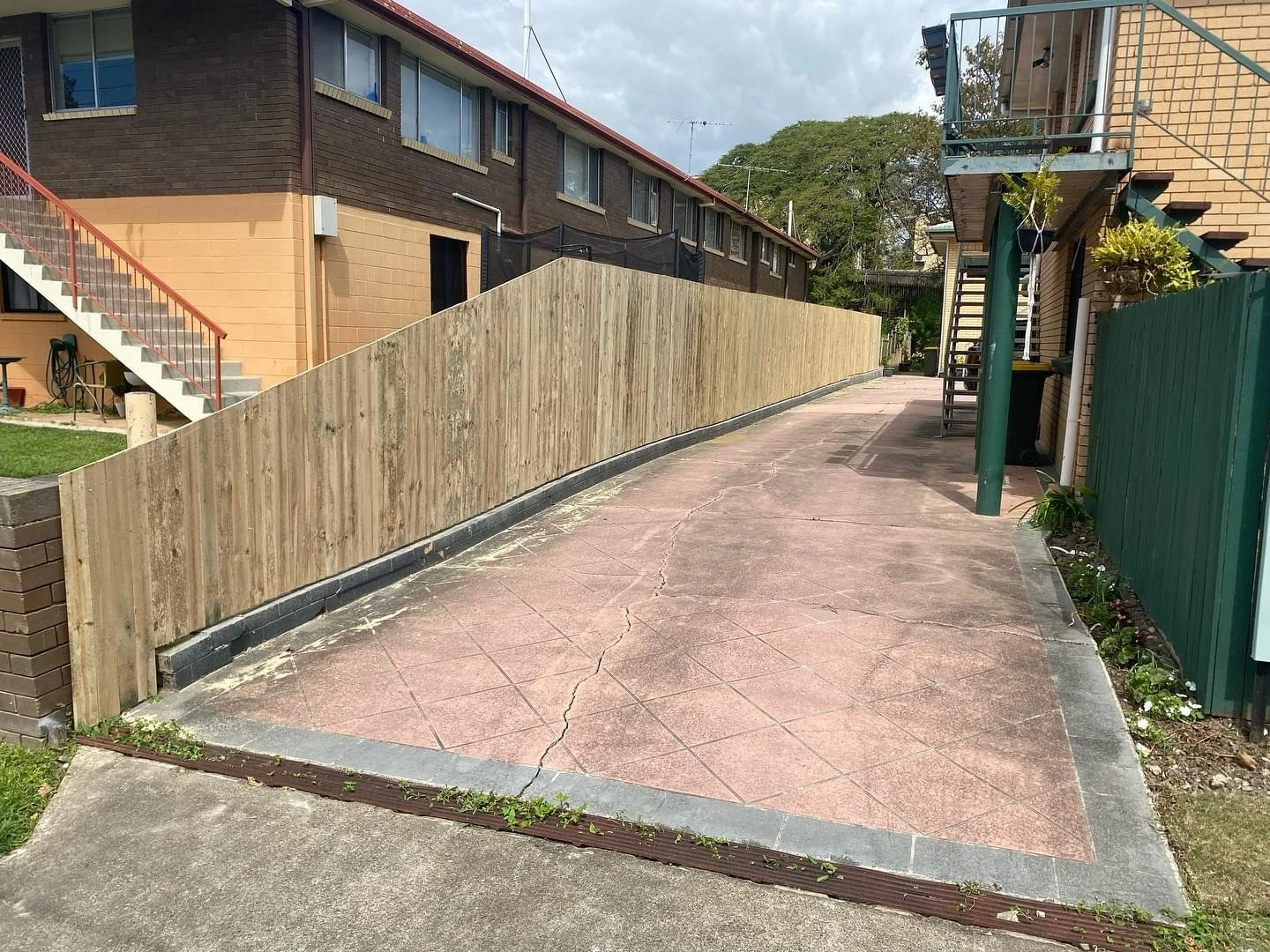A concrete walkway with visible cracks, bordered by a wooden fence on the left and a green metal fence on the right, with residential buildings and outdoor stairs visible in the background.