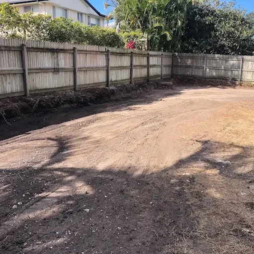 Empty backyard with dirt ground, wooden fence, and some trees and plants in the background.