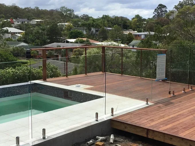 View of a rooftop pool and wooden deck with a glass railing, overlooking a residential neighborhood with trees and houses under a cloudy sky.