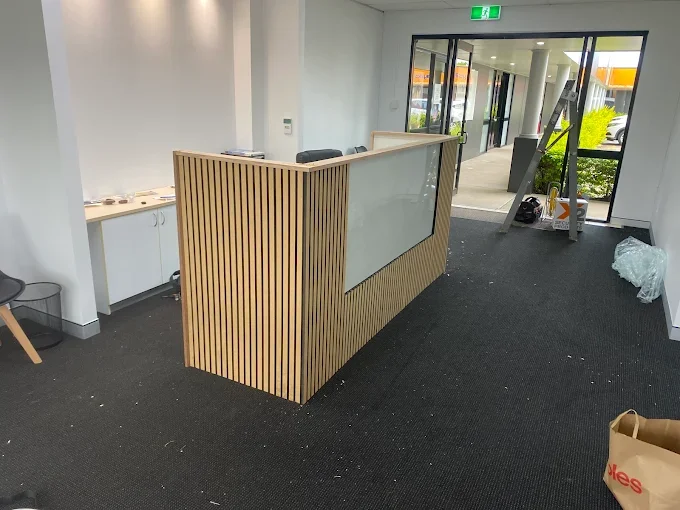 Empty reception area with a modern wooden and glass counter, black carpet, and a glass door leading outside with greenery visible.