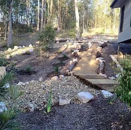 A garden path made of stepping stones with a wooden border, surrounded by dirt, rocks, and some new plants, next to a house with a grey exterior.