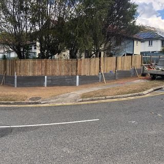 A construction site with a wooden fence surrounding it. The fence is supported by metal poles, and there is a boat parked nearby on the street. In the background, there are trees and residential buildings under a partly cloudy sky.