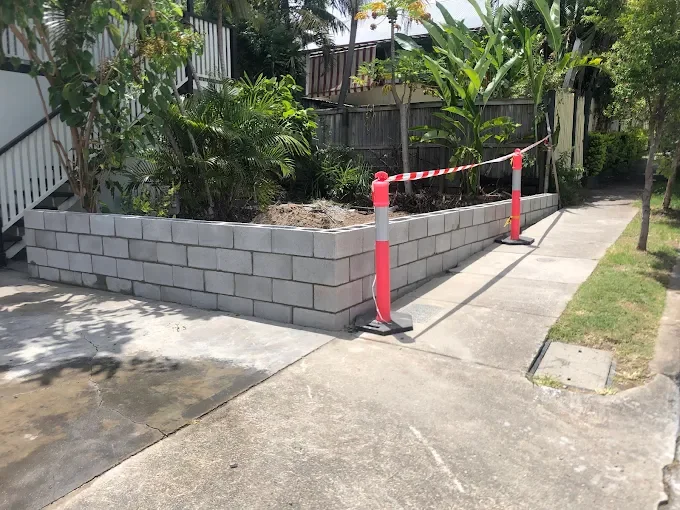 A sidewalk with a new concrete block garden bed surrounded by trees and plants, with construction cones and tape indicating ongoing work.