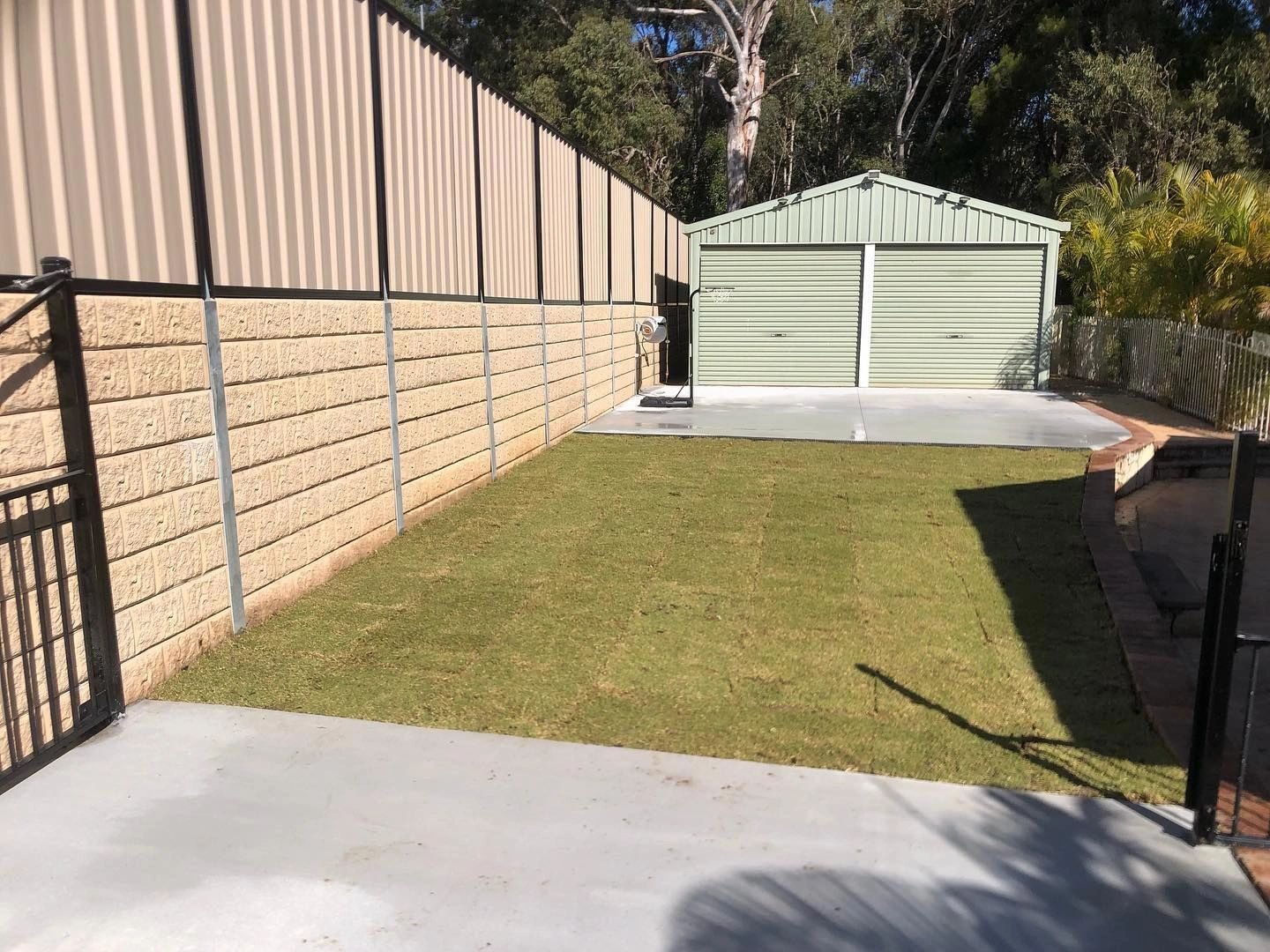 A backyard with a small grass area, a concrete patio, and a light green garage with closed roller doors. The yard is enclosed by a beige brick wall and a black metal fence, with trees in the background.