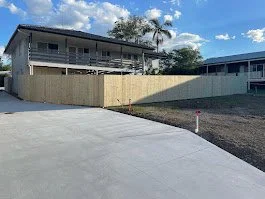 View of a residential area with two houses, a wooden fence, a concrete driveway, and a clear blue sky with some clouds.