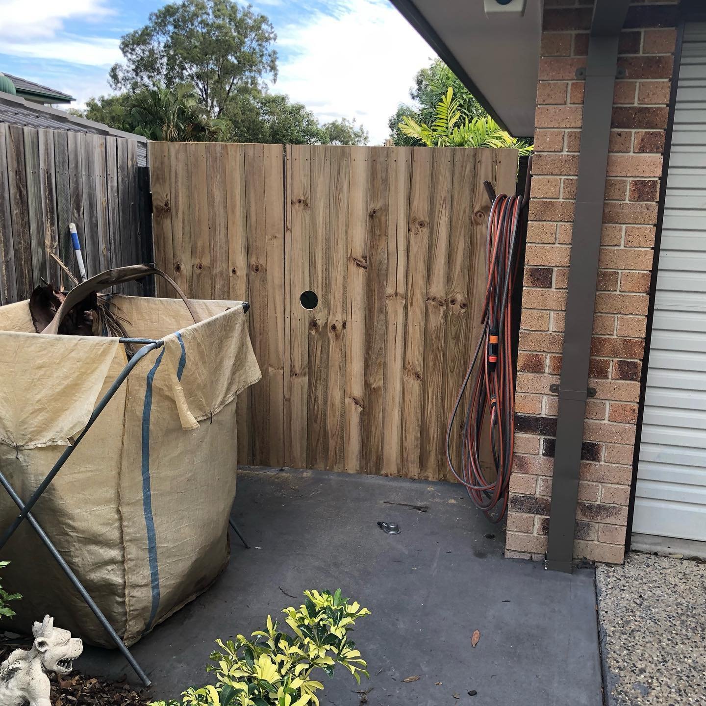 Partial view of a backyard with a wooden fence, brick wall with hanging hoses, a laundry tub, and some greenery.