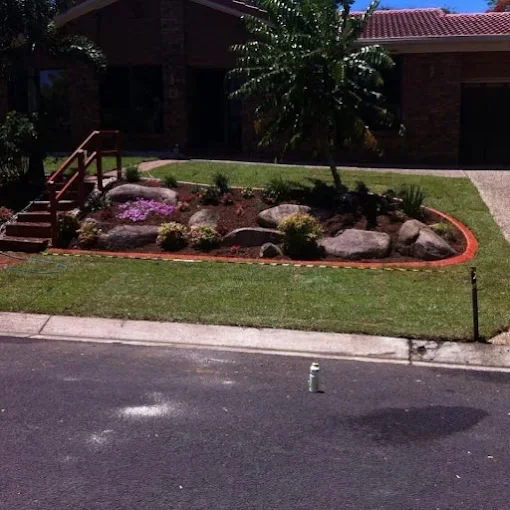 A landscaped front yard with a small garden bed containing rocks, plants, and a tree, bordered by red brick edging, in front of a house with a brick exterior and stairs leading up to the porch.