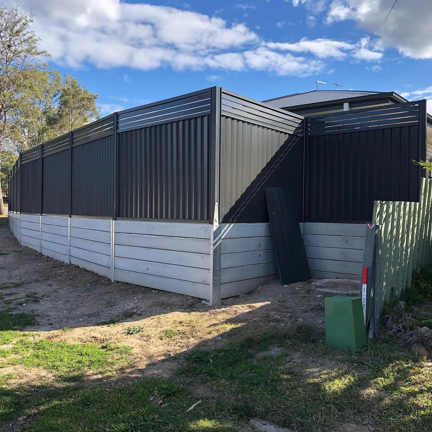 A newly installed black and grey privacy fence surrounds a yard with a house in the background, underneath a partly cloudy blue sky.