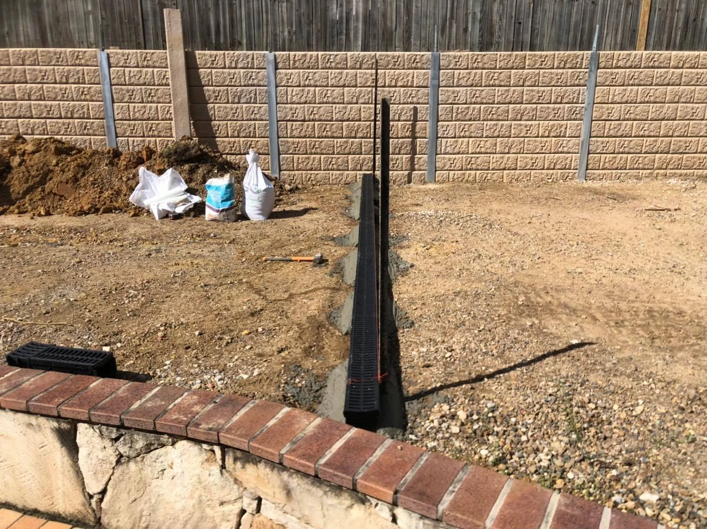 A construction site shows a gravel yard with a long black drainage or utility trench in the center, surrounded by dirt, with a brick wall at the foreground and a beige brick fence at the background. Construction materials and tools are visible, including white sandbags, a loose hammer, and a plastic drain cover.