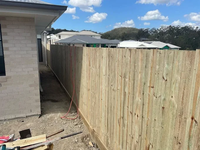 New wooden fence under construction along the side of a house with a dirt ground in a residential area.