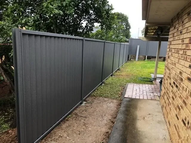 A gray metal privacy fence along a backyard with a brick house, a concrete walkway, and green grass and trees in the background.
