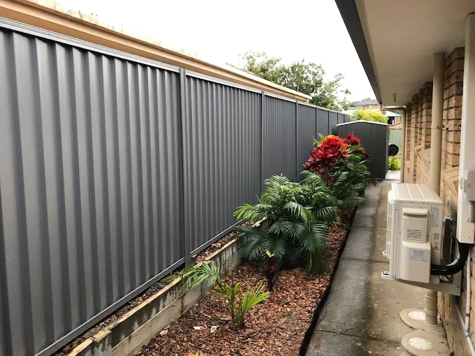 A narrow backyard garden bed with tropical plants and a red-leafed shrub, bordered by a gray metal fence and a sidewalk on the right side, next to a brick house with visible air conditioning units.