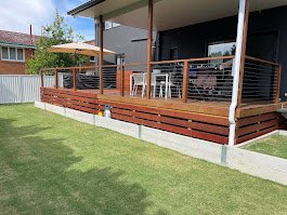 Backyard deck with a wooden railing and furniture, attached to a modern house with large windows and a patio umbrella, surrounded by a green lawn.