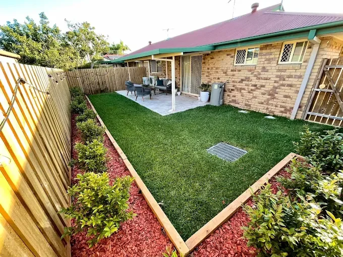 Backyard with a grassy lawn, bordered by a wood fence and flowerbeds filled with shrubs and mulch, and a patio area with outdoor furniture.