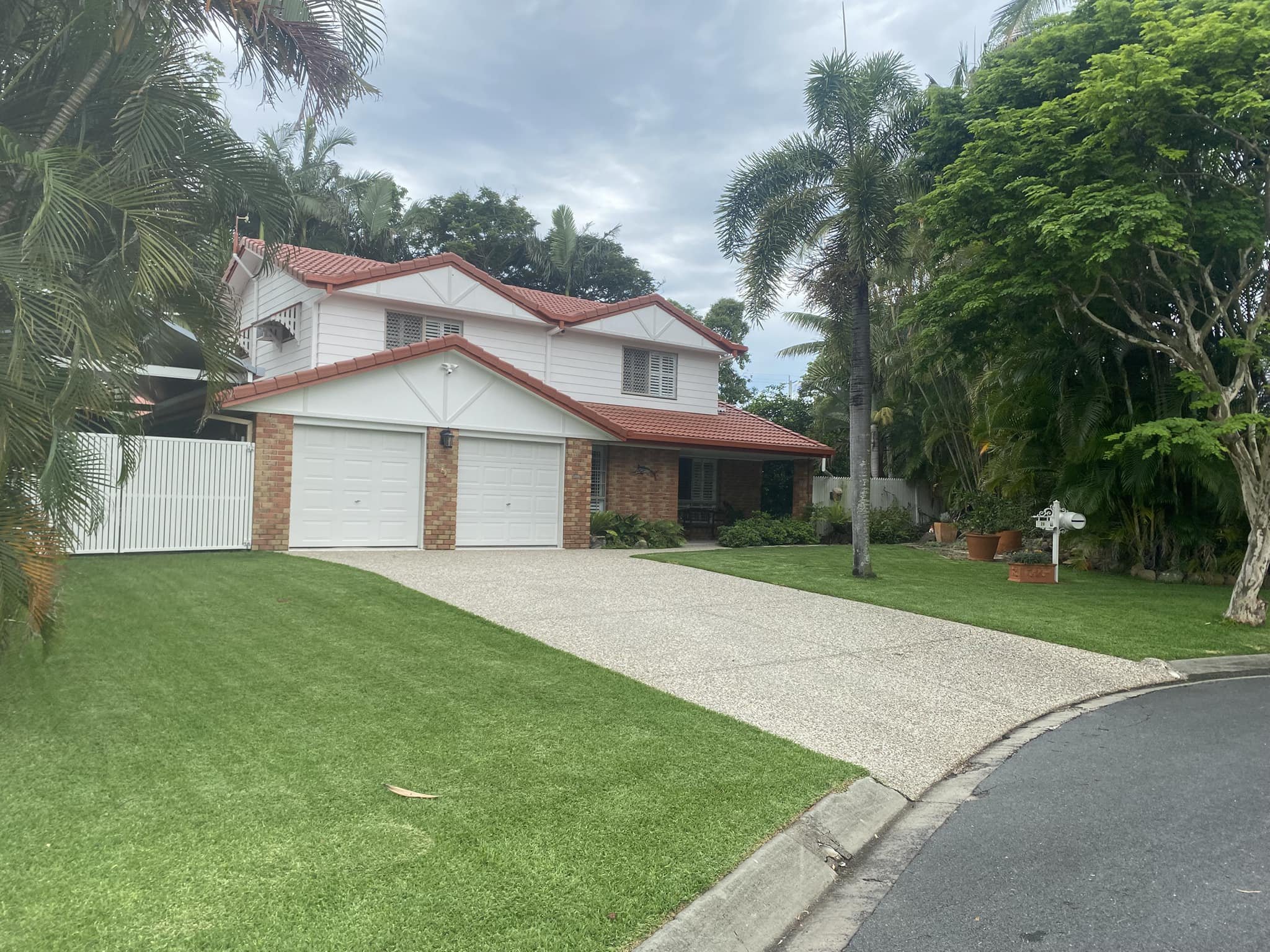A two-story house with white walls, a red tiled roof, and brick accents on the garage. The house is surrounded by green trees and manicured lawn, with a gravel driveway leading up to the garage.