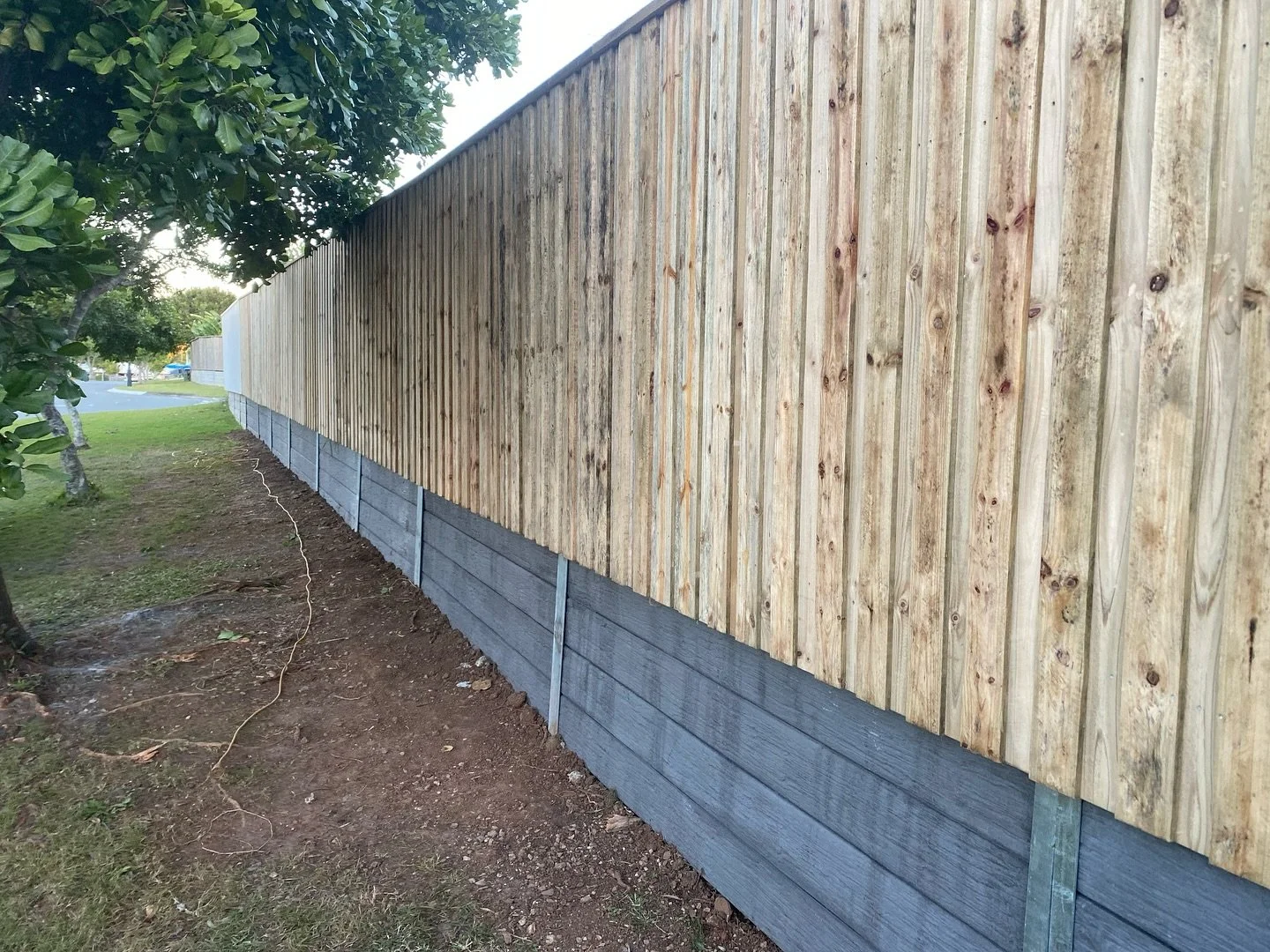 A newly constructed wooden fence with vertical planks and a concrete base, located next to a grassy yard with trees.