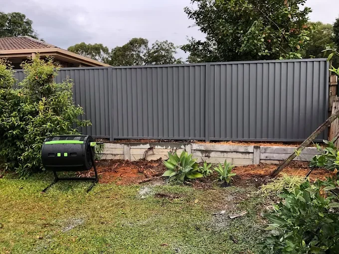 A backyard with a new gray metal fence, some plants, and a black compost bin on the grass.