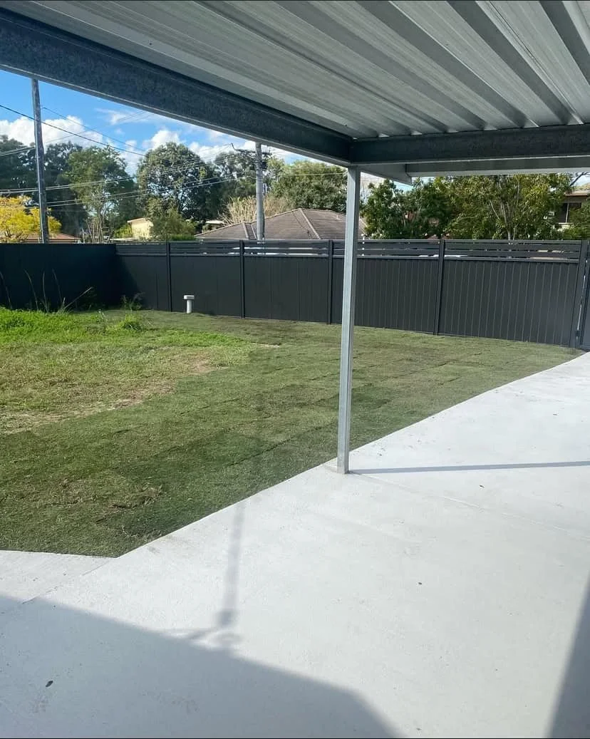Backyard patio with a covered area, green grass, black privacy fence, trees, and neighboring houses in the background.