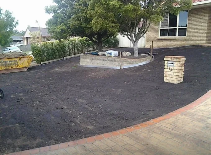A front yard with freshly tilled dark soil, two trees, a brick mailbox, and a raised garden bed in a suburban neighborhood.