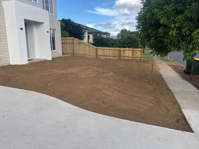 A residential front yard with freshly tilled soil, a concrete sidewalk on the right, a white modern house on the left, a green tree, and a wooden fence in the background under a partly cloudy sky.