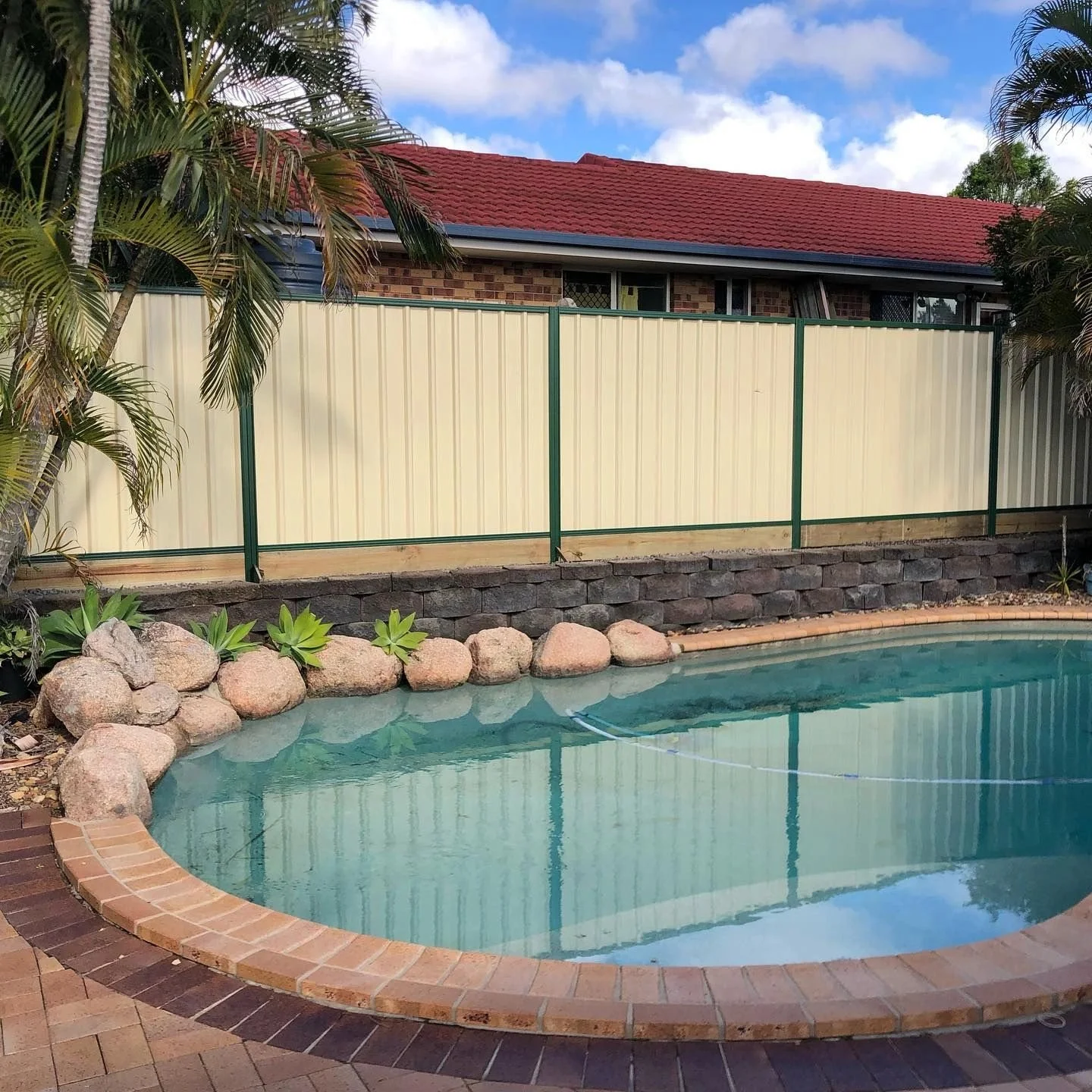 Swimming pool with brick border, surrounded by plants, rocks, and a beige fence, with a house and blue sky in the background.