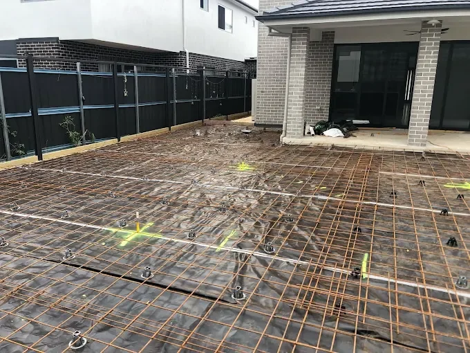 Construction site showing a concrete slab with rebar reinforcement for future paving or concrete pouring, adjacent to a house with a screened patio, in a residential backyard.