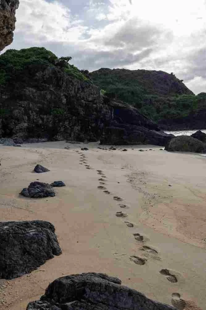 Footprints in the sand on a beach with rocks and green hills under a cloudy sky.