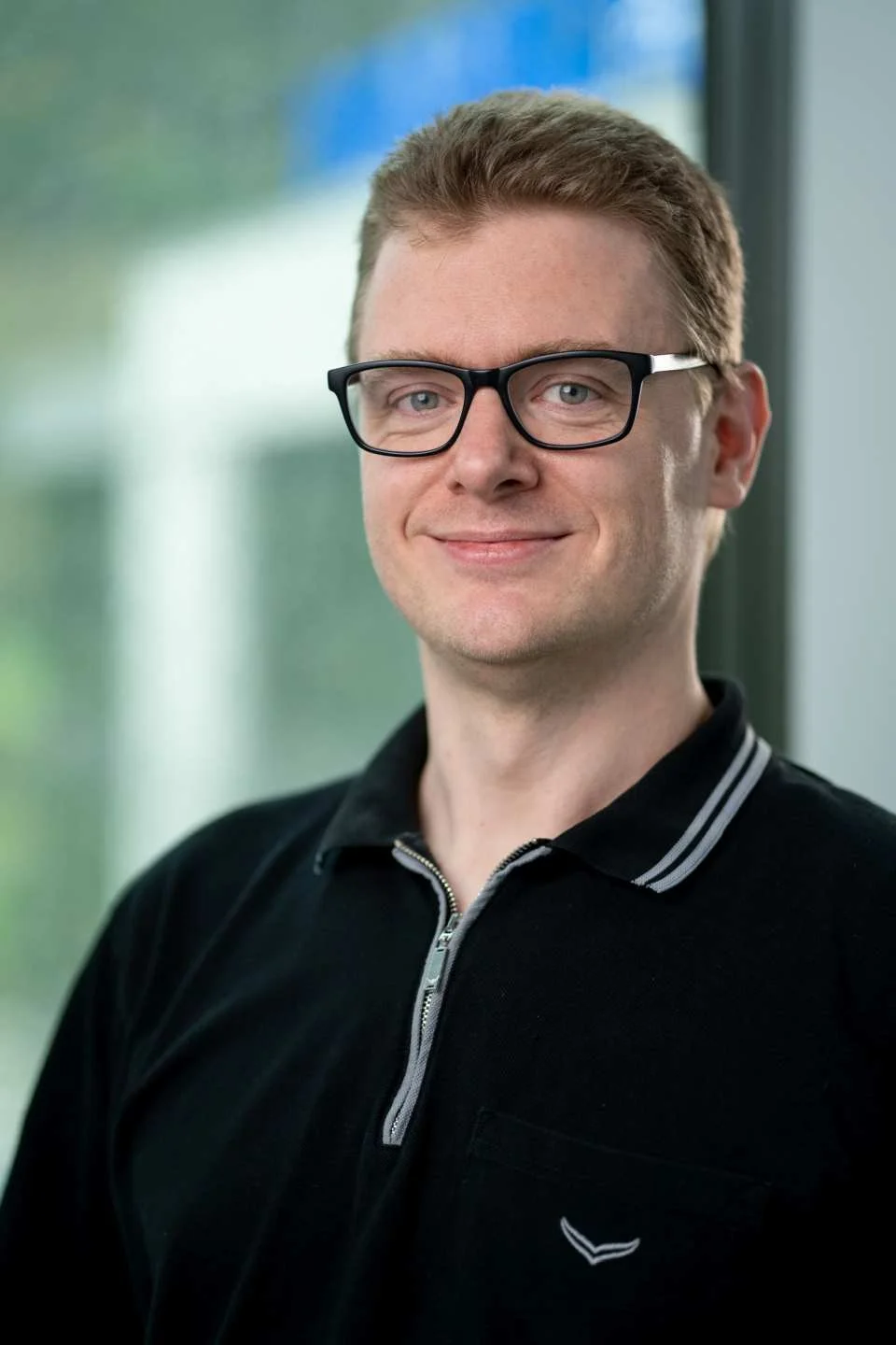 A man with short brown hair, glasses, and light skin, smiling and standing indoors near a window with greenery outside.